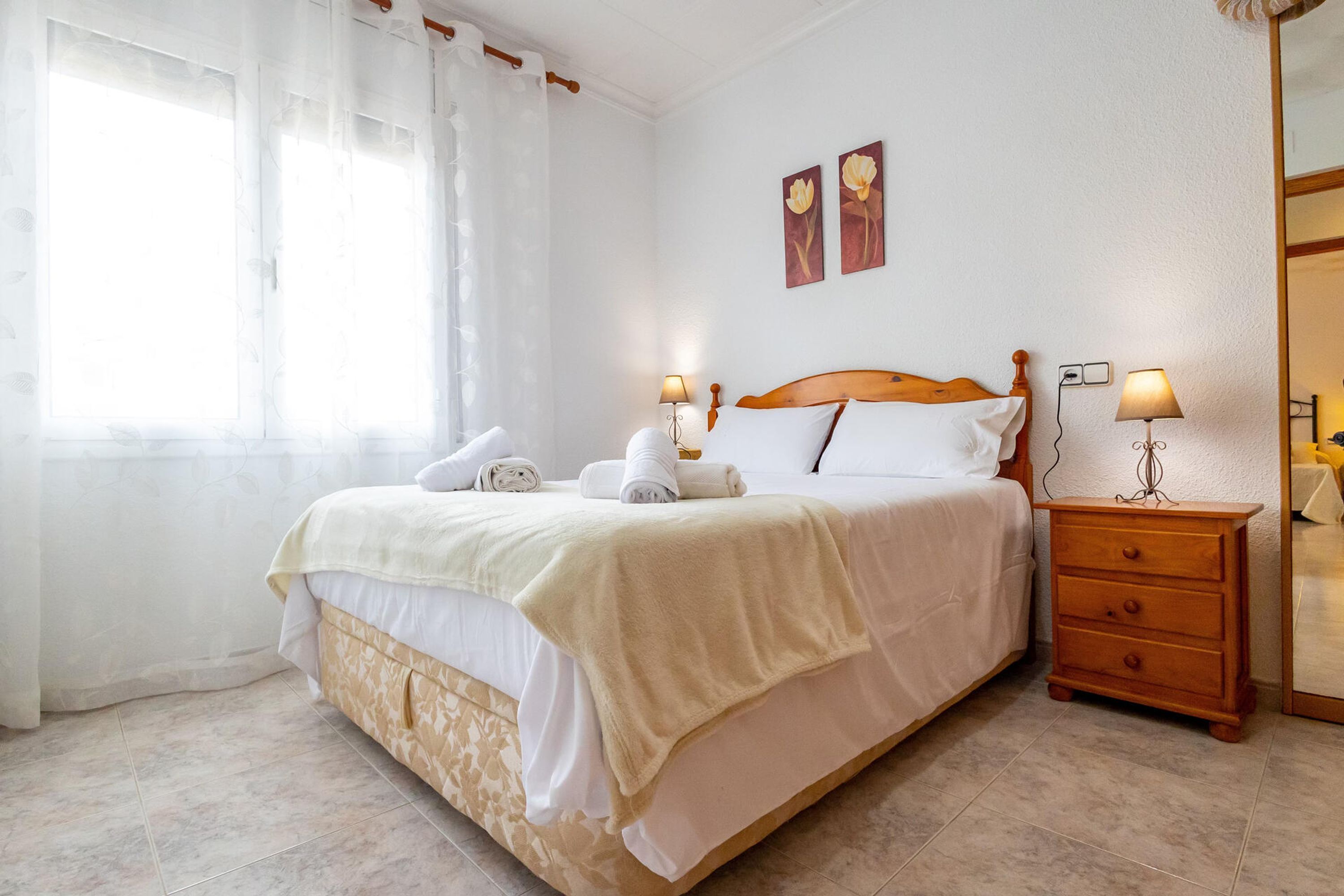 Un lit bien fait, avec du linge de lit blanc et une couverture beige, une table de chevet en bois avec deux lampes, et une fenêtre ornée de rideaux légers.