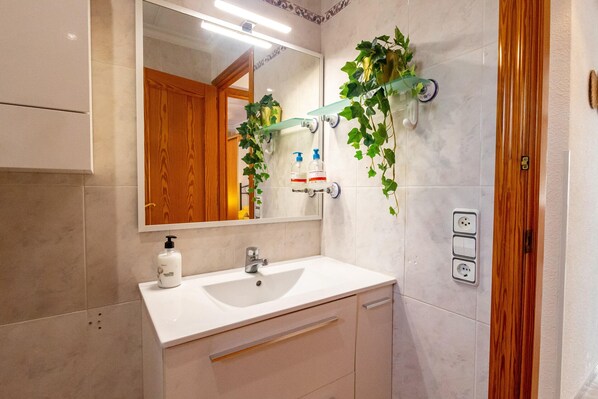 A bathroom with a white sink, mirror, and wooden door. There are plants on the wall and a shelf with toiletries.
