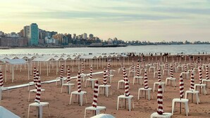Una playa con hileras de sillas blancas y sombrillas a rayas, un horizonte urbano al fondo y un mar tranquilo.