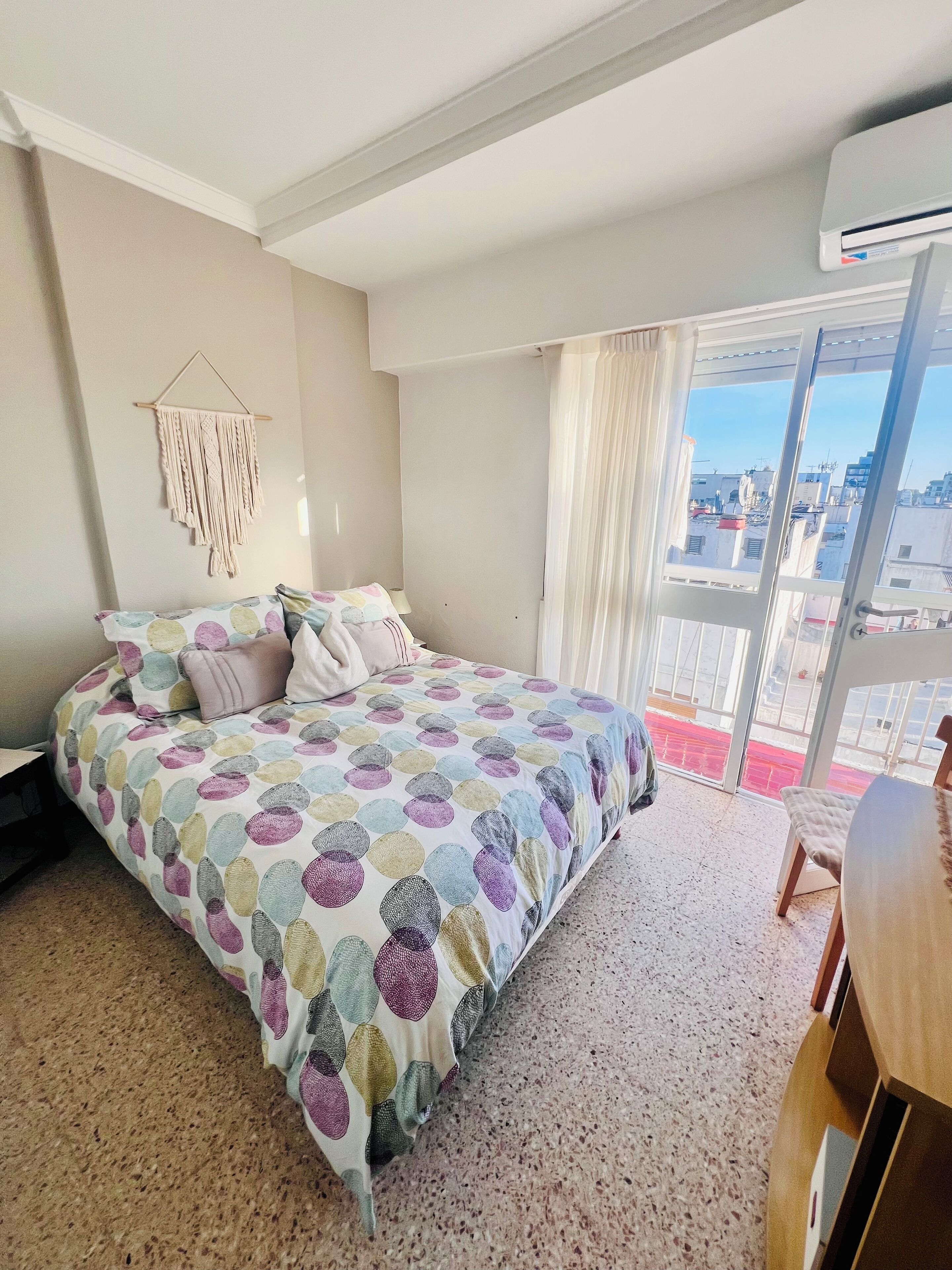 Un dormitorio con cama, un colgador de macramé, una ventana con cortinas y vistas a edificios.