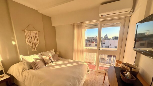 A hotel room with a bed, a desk, a TV, and a balcony with a city view.