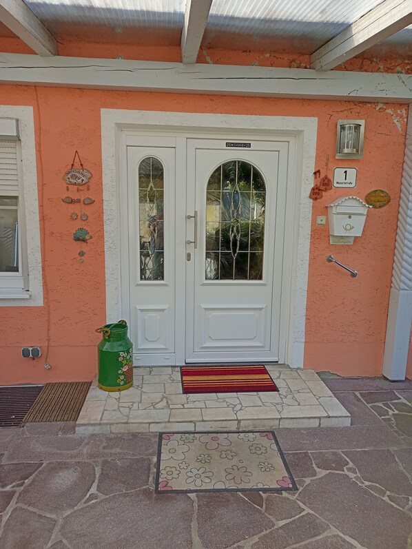 Une maison avec une entrée à deux portes, une bouteille de gaz verte, un paillasson rouge et jaune, et une allée d’entrée pavée de pierres.