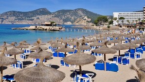 A beach with rows of blue lounge chairs and thatched umbrellas, a rocky coastline, and a building with balconies overlooking the sea. - Port Andratx Family Holiday House With Pool (Andraitx)