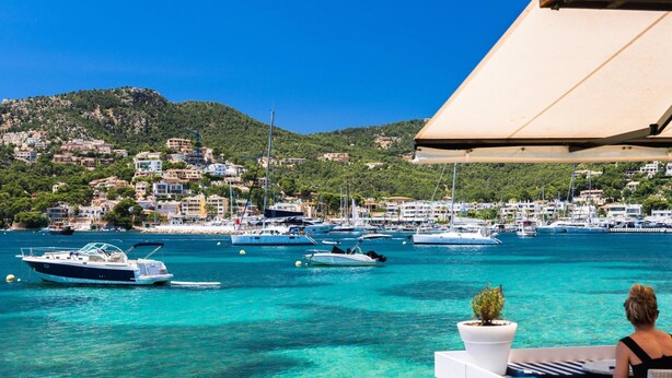 A coastal view with boats, a woman on a balcony, and a hillside town. - Port Andratx Family Holiday House With Pool (Andraitx)