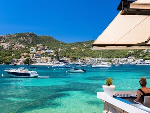 Panorama sulla costa con barche, una donna su un balcone e una cittadina collinare.