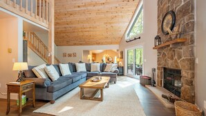 A spacious living room with a wooden ceiling, a stone fireplace, and a large window providing natural light.
