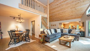 A spacious living room with a wooden ceiling, a dining area with a round table and chairs, and a large gray sofa with multiple pillows.