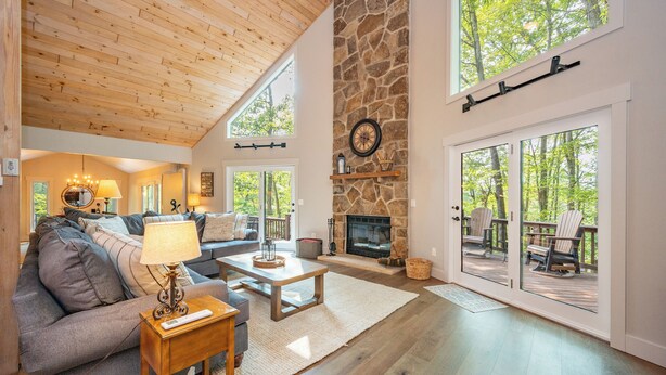 A spacious living room with a stone fireplace, wooden ceiling, and large windows offering a view of the outdoors.