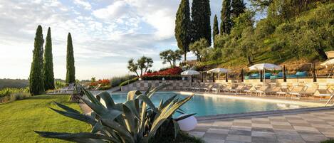 A pool area with lounge chairs and umbrellas, surrounded by greenery and tall trees.