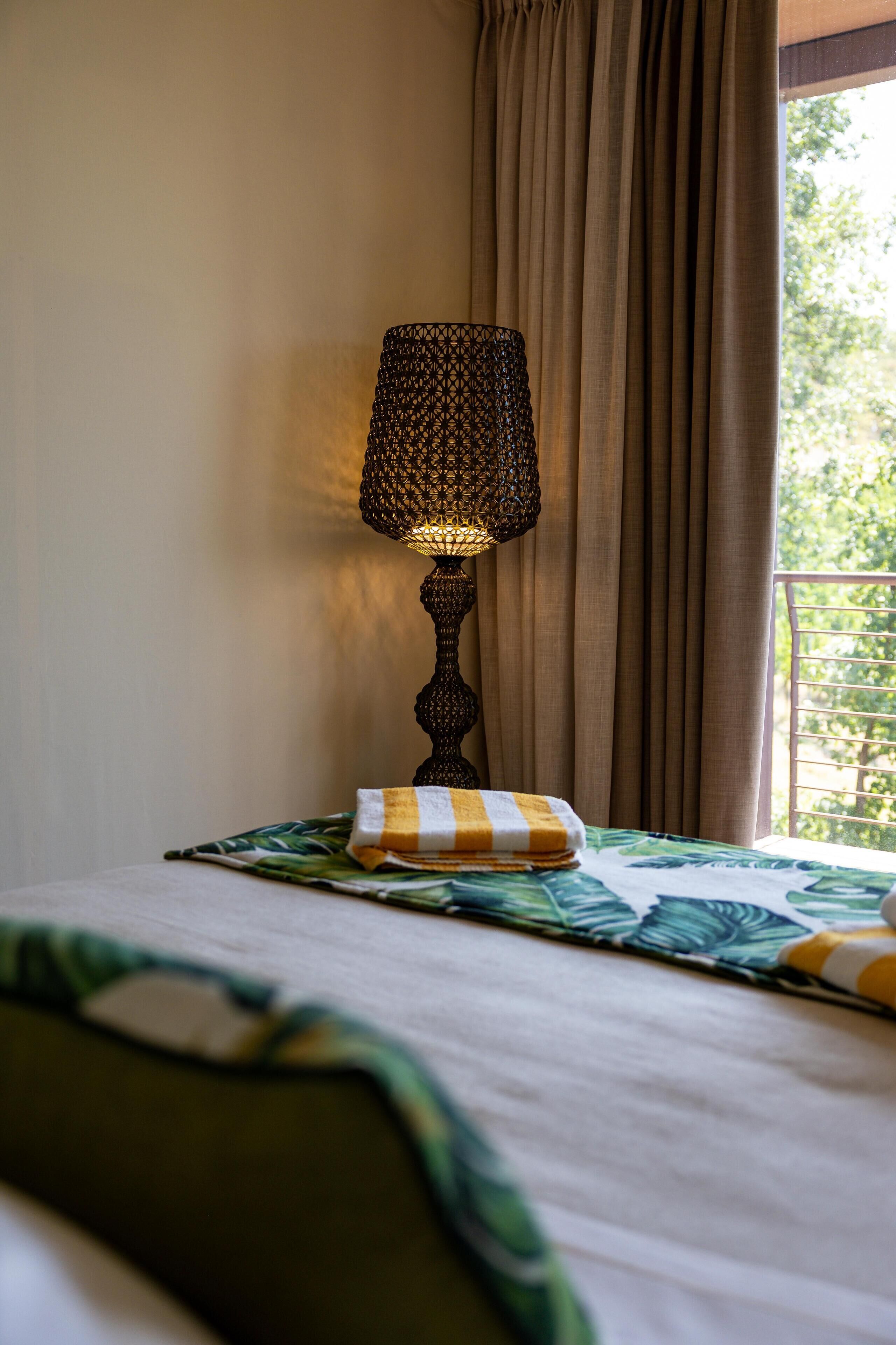 Un lit avec une couvre-lit à motifs de feuilles vertes, un coussin rayé et une lampe de table à motifs foncés.