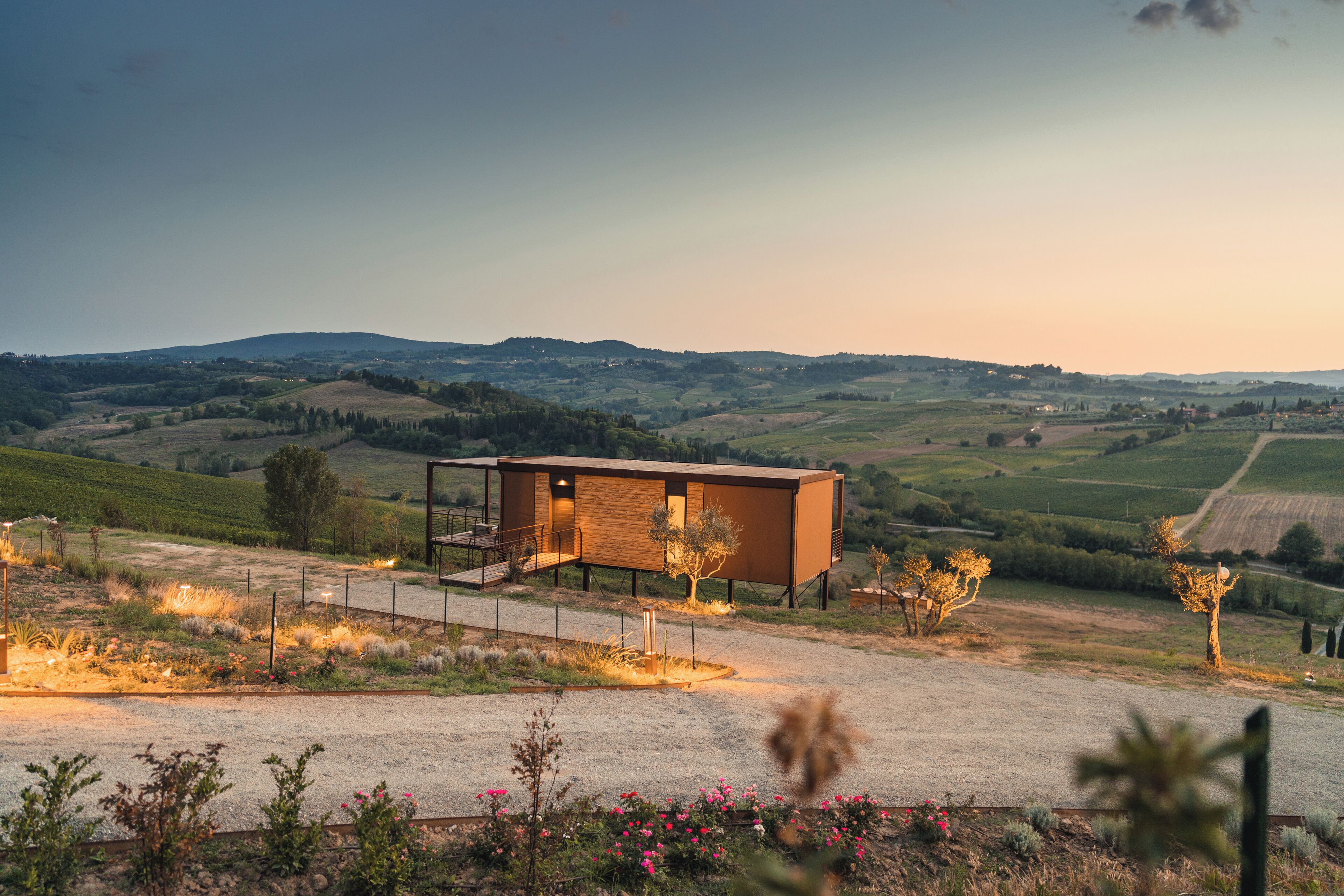 A modern house with a wooden exterior and a glass entrance, surrounded by a gravel driveway and flowering plants.