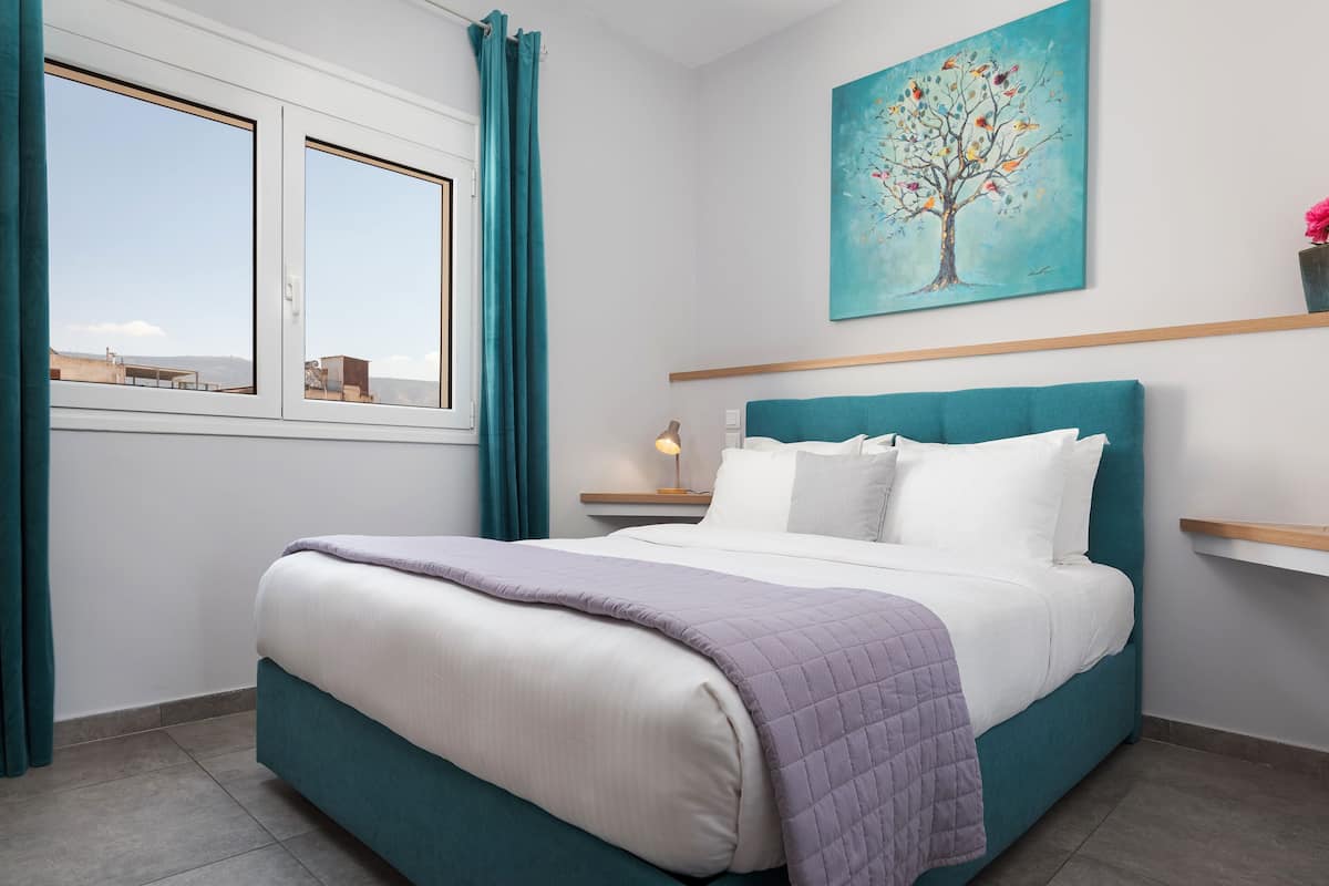 A bedroom with a bed, a nightstand, a lamp, a painting, and a window with curtains.