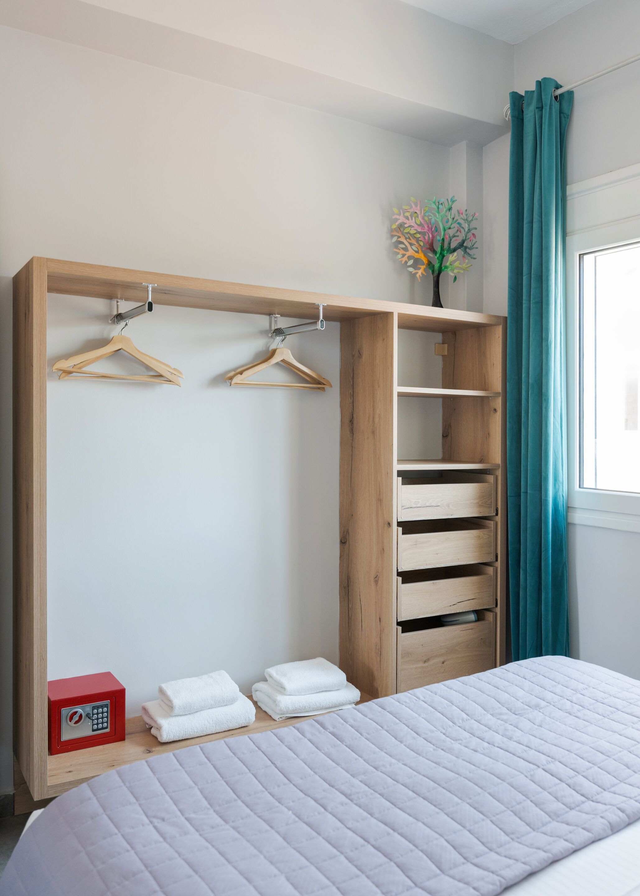 A bedroom with a bed, a wooden wardrobe with hanging space, a small safe, and a window with curtains.