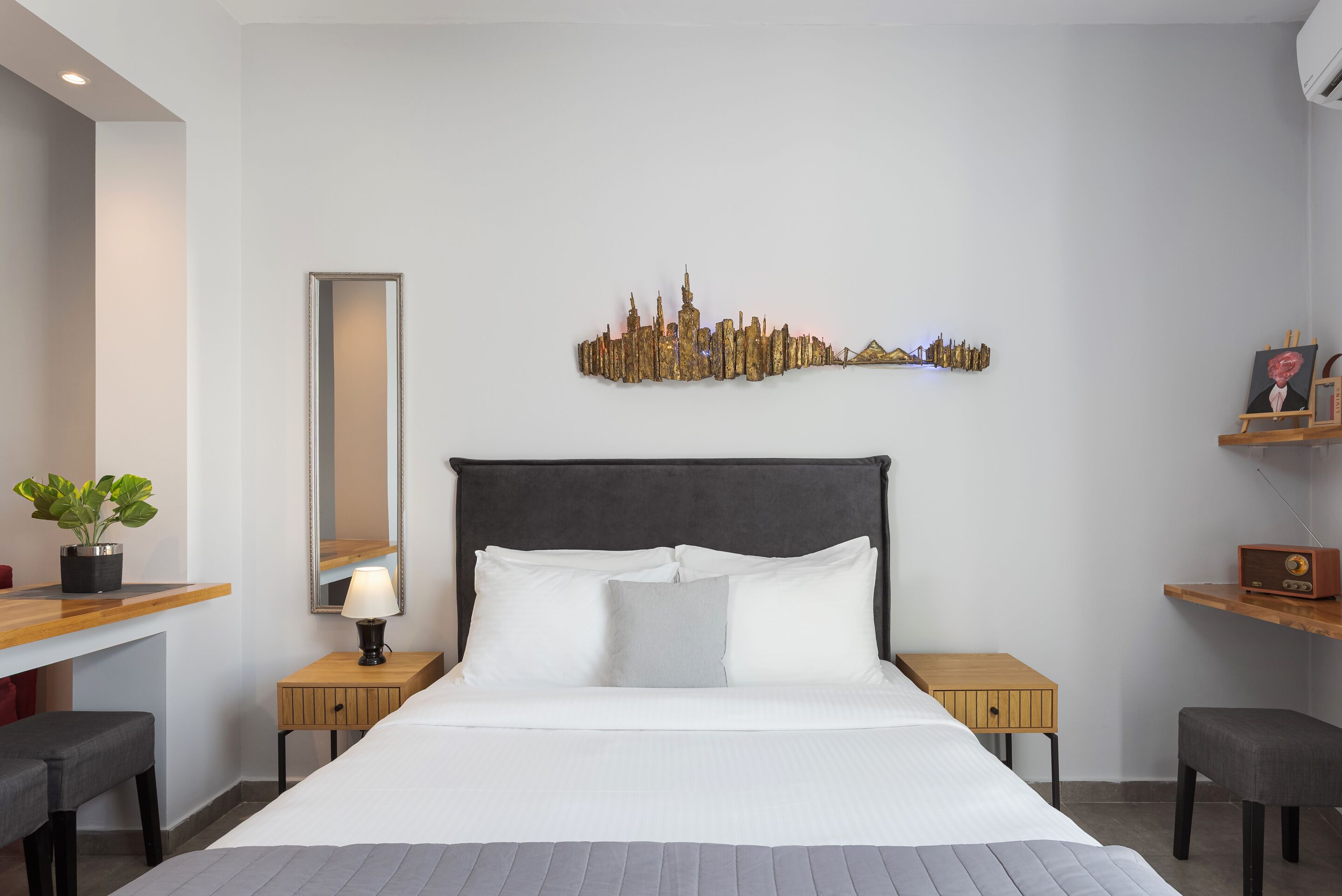 Une chambre d’hôtel moderne avec un grand lit, une œuvre d’art représentant un paysage urbain accrochée au mur, une table de chevet avec une lampe, une petite plante en pot et une étagère en bois avec une radio.