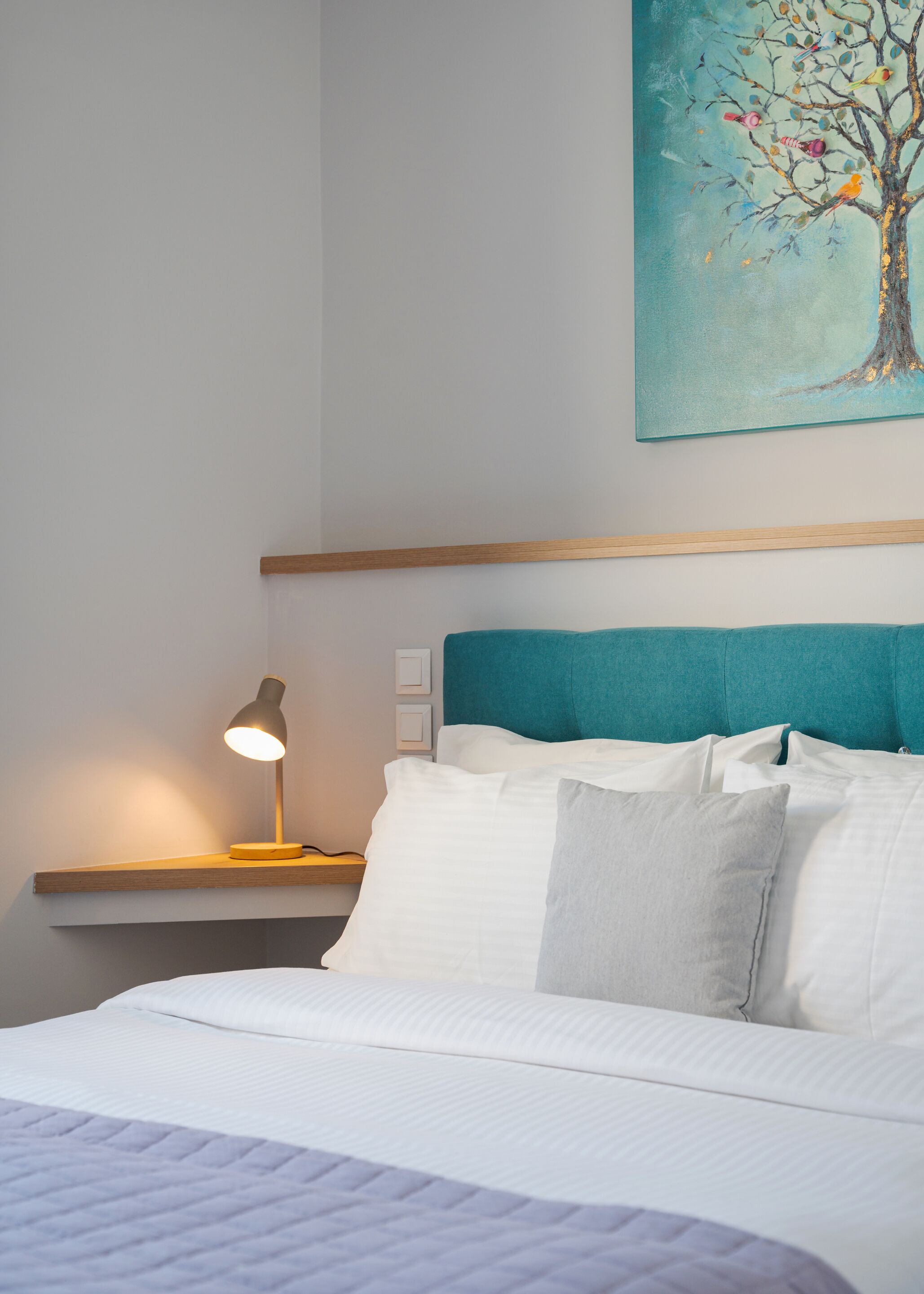 Cama bem arrumada com lençóis brancos e cabeceira verde-água. Um criado-mudo com luminária é visível.