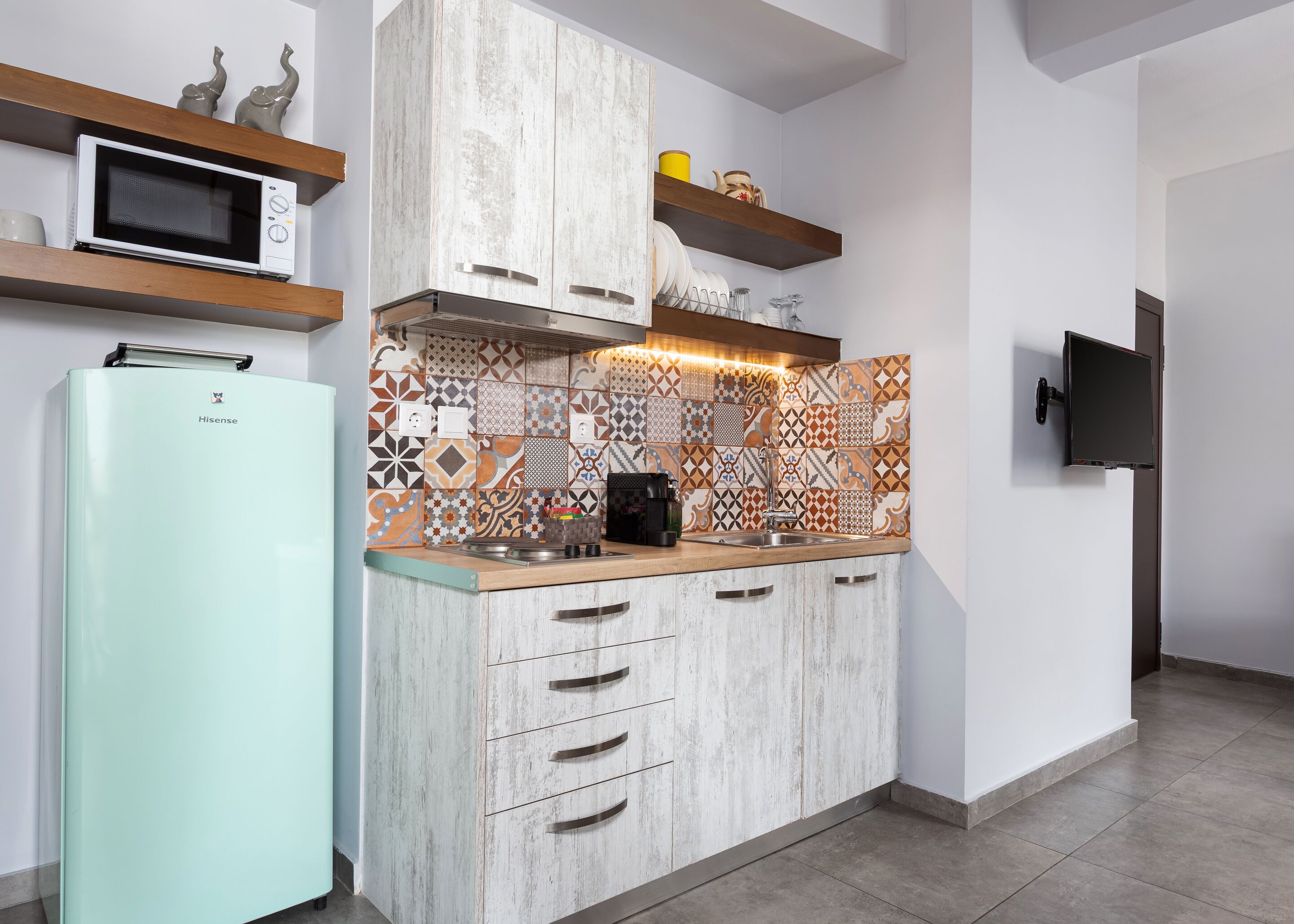 A modern kitchen with a Hisense refrigerator, wooden cabinets, a patterned backsplash, and a flat-screen TV mounted on the wall.