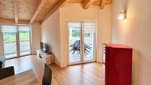 Ein geräumiges Zimmer mit Holzbalkendecke, einem Fernseher auf einem Standfuß, einem roten Schrank und einer Glastür, die zu einem Außenbereich mit Sitzgelegenheiten führt.