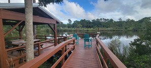 A wooden deck with picnic tables and chairs overlooking a calm lake, surrounded by dense forest. - 2-BR Getaway w/ HUGE screened porch, Golf Cart, Pool, & Boat Ramp near Wolf Bay (Elberta)