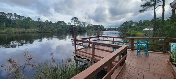 A wooden deck with a railing overlooking a calm river, surrounded by dense greenery. - 2-BR Getaway w/ HUGE screened porch, Golf Cart, Pool, & Boat Ramp near Wolf Bay (Elberta)