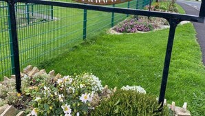 A wooden sign with text in a foreign language, surrounded by a green fence and a flower bed.