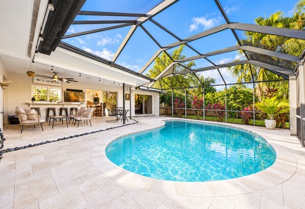 A modern indoor swimming pool with a glass ceiling, surrounded by a patio area with seating and a bar.