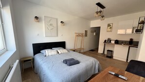 Un dormitori modern amb un llit gran, taules de nit, una taula de nit de fusta, una cadira de fusta, una taula petita amb un comandament a distància, una zona de cuina amb un rentapeus, fogó i armaris, i un quadre amb marc a la paret.