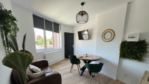 A modern living room with a sofa, a dining table with chairs, a TV, and a large window with blinds. - Le Jardin Botanique : Balnéo 2p (Lens)