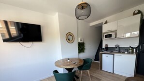A modern kitchen with white cabinets, a microwave, and a coffee machine. There is a dining area with a round wooden table and two green chairs. - Le Jardin Botanique : Balnéo 2p (Lens)