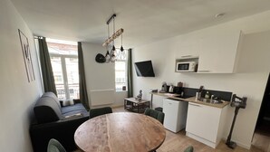 A compact living space with a dining table, green upholstered chairs, a sofa, a kitchen area with a microwave, and a TV mounted on the wall.