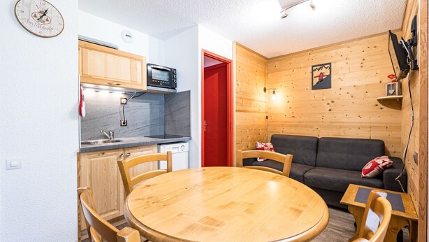 A compact kitchen with wooden cabinets, a microwave, and a sink. There is a dining table with chairs and a sofa in a living area with wooden walls.