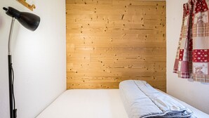 A small, neatly made room with a wooden wall, a single bed, a lamp, and a patterned curtain.