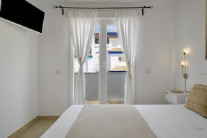 A bedroom with a bed, white curtains, a nightstand, a wall-mounted lamp, and a view of buildings outside.