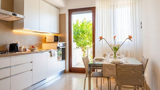 Une cuisine moderne avec des meubles blancs, un coin repas doté d’une table en verre et une vue sur la verdure par une porte.