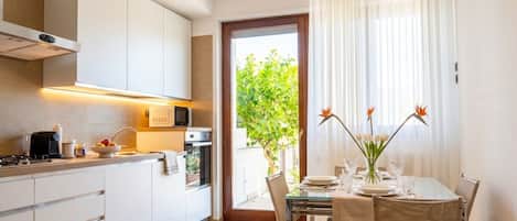 A modern kitchen with white cabinets, a dining area with a glass table, and a view of greenery through a door.