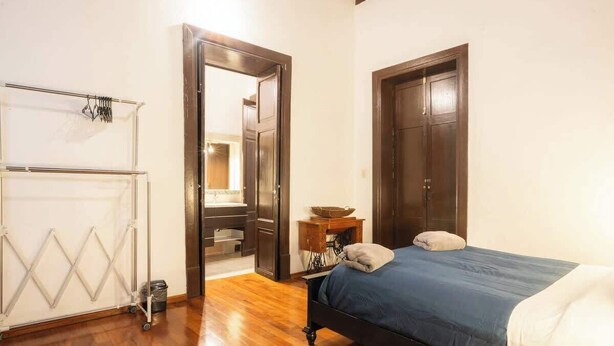 A bedroom with a wooden floor, a bed with a blue bedspread, a wooden wardrobe, and a white radiator. - Regina 70 · Amazing Historic Colonial Duplex by Cathedral (Mexico City)