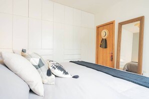Un lit bien fait, avec des oreillers, une porte en bois sur laquelle est accroché un chapeau, et un miroir.