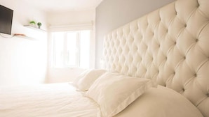 Ein Schlafzimmer mit einer gepolsterten Kopfstütze, einem an der Wand montierten Flachbildfernseher und einem Fenster, das für natürliches Licht sorgt.