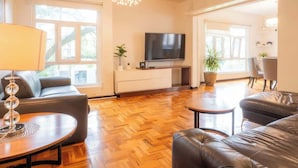 Ein geräumiges Wohnzimmer mit Flachbildfernseher, Ledersofas, Holzboden und großen Fenstern.