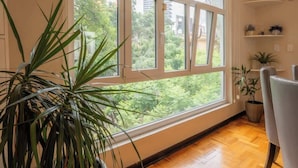 Ein Zimmer mit großem Fenster, einem Blumentopf, einem Sessel und einem Regal mit Pflanzen.