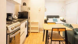 Une cuisine compacte équipée d’une cuisinière, d’un évier et de meubles blancs. On y trouve également un coin repas avec une table et des chaises, et un lit avec une tête de lit en arrière-plan.