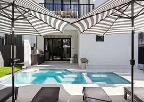 Un área de piscina exterior moderna con sombrillas a rayas, sillas de descanso y una zona de barbacoa.