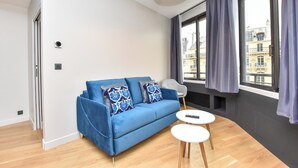 A modern living room with a blue sofa, two white side tables, and a large window with curtains. - Chic Apartment - 1br/4p - Near Opera Garnier (Paris)