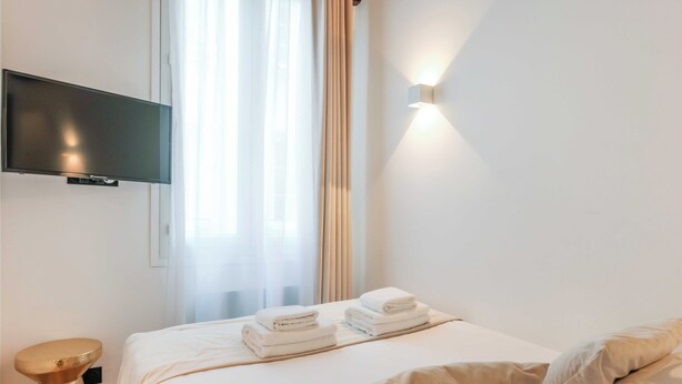 A hotel room with a bed, a flat-screen TV mounted on the wall, a bedside table, and a wall-mounted light fixture. - Relaxing Urban Getaway, Notre Dame (Paris)