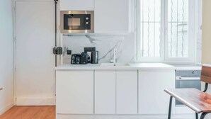 A modern kitchen with white cabinetry, a stainless steel microwave, a coffee maker, and a window with blinds. - Relaxing Urban Getaway, Notre Dame (Paris)