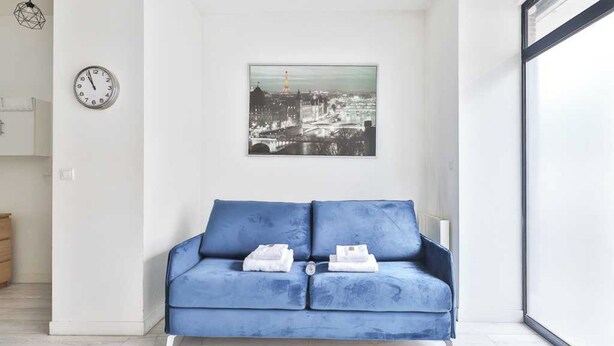 A modern living room with a blue sofa, a cityscape painting, a clock, and a minimalist decor. - Bright Apartment - 1br/4p - Bibliotheque Francois Mitterrand (Paris)