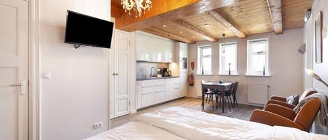 A bedroom with a large bed, a TV mounted on the wall, a dining area with a table and chairs, and a kitchen in the background.