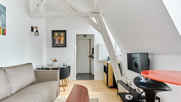 A modern loft apartment with a high ceiling, a dining area with a black table and chairs, a kitchen with a white refrigerator, and a TV on a stand. - Charming 1 Bedroom Flat Heart of Marais (Paris)