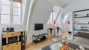 A modern loft-style room with a loft bed, a dining area with a table and chairs, a kitchen area with a microwave and coffee maker, and a television on a wall. - Charming 1 Bedroom Flat Heart of Marais (Paris)
