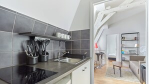 A modern kitchen with a black countertop, stainless steel sink, and a built-in oven. There is a dining area with a sofa and a small table. - Charming 1 Bedroom Flat Heart of Marais (Paris)