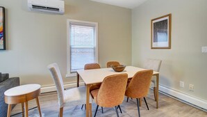 Un comedor con una mesa, sillas y un tazón. Hay una ventana con persianas, un equipo de aire acondicionado y un cuadro en la pared.